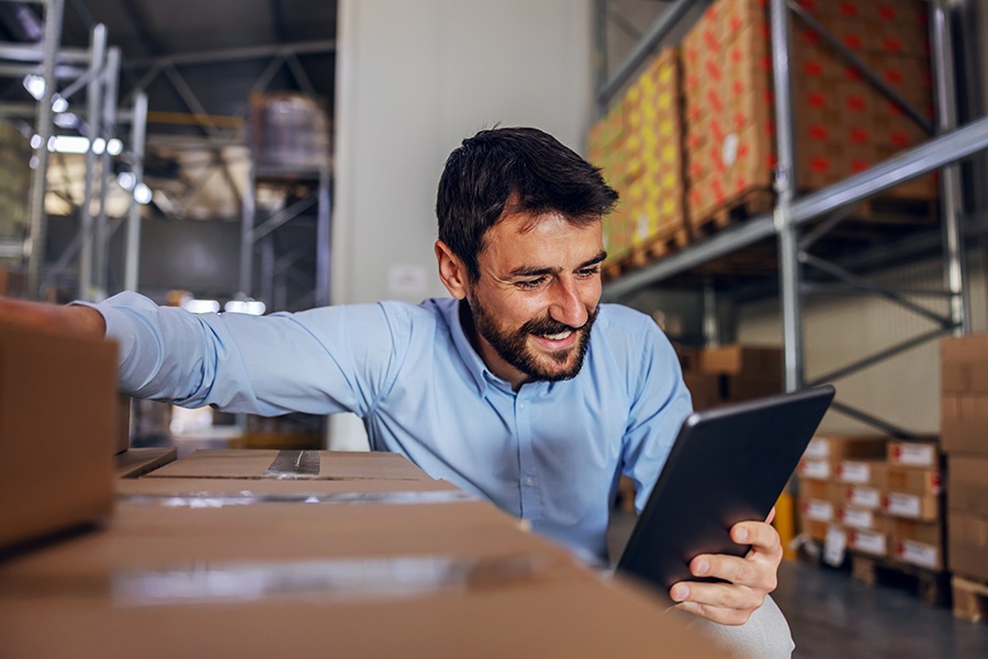 Blog - Portrait of a Cheerful Young Male Business Owner Using a Tablet While Crouching Next to Boxes Inside a Warehouse Facility